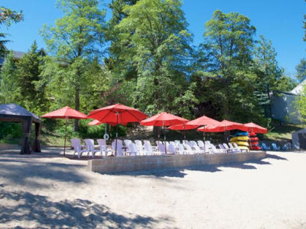 Sandy beach with red umbrellas, white chairs, and a gazebo amidst green trees.