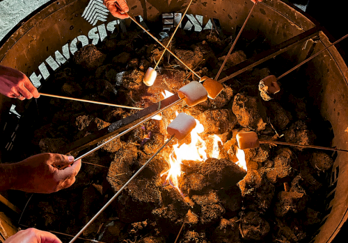 Gloved hands roast marshmallows on long sticks over an open fire in a large metal grill.
