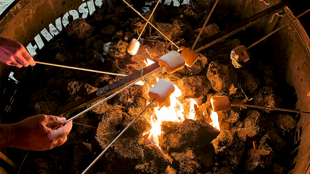 Gloved hands roast marshmallows on long sticks over an open fire in a large metal grill.
