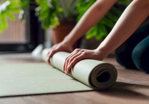 Hands rolling up a yoga mat on a wooden floor, with greenery in the background.