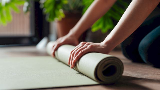 Hands rolling up a yoga mat on a wooden floor, with greenery in the background.