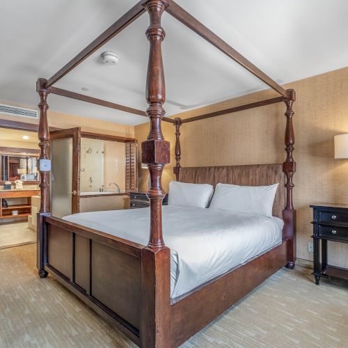 A hotel bedroom with a four-poster bed, white bedding, nightstands, a lamp, red curtains, and a bathroom in the background.