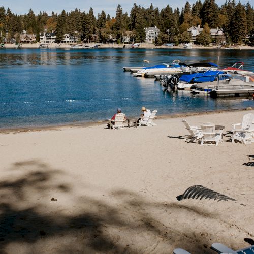 A sunny lakeside beach with white chairs, anchored boats at a dock, and homes backed by tall pine trees.