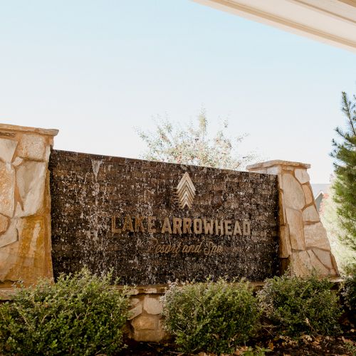 A stone &ldquo;Lake Arrowhead&rdquo; sign with a cascading water feature and trees in the background under a bright sky.