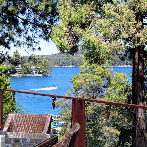 Lakeside view through pine trees: blue water with a passing boat and distant shoreline, seen from a balcony patio.