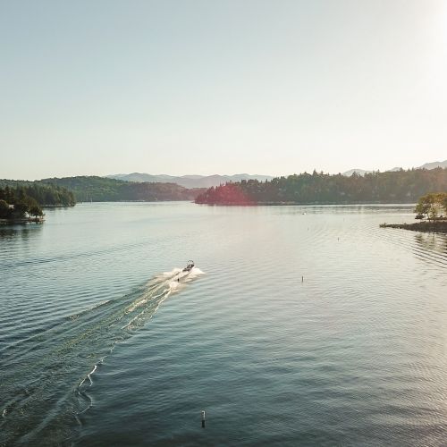 A wide lake at sunset with forested hills, a boat speeding across, and sunlit shoreline buildings.