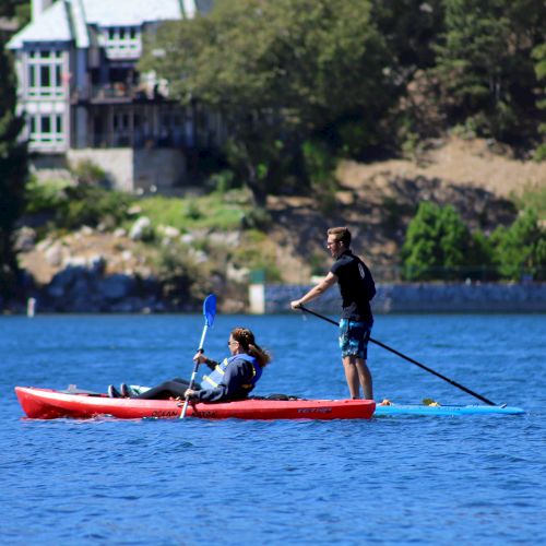 Two people paddle a red kayak on bright blue water near a leafy shoreline with houses in the background.
