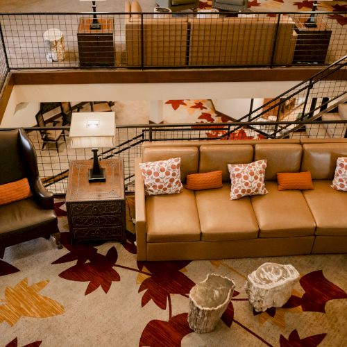A cozy living room loft with a beige sectional sofa, patterned pillows, coffee table, and a metal-railed upper balcony above.