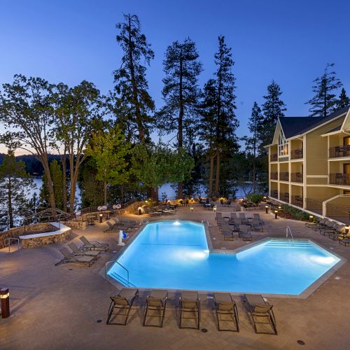 A lakeside hotel courtyard at dusk with a glowing swimming pool, lounge chairs, and multi-story rooms overlooking the water.