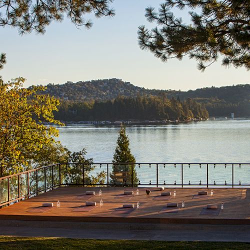 A lakeside outdoor deck set for a meeting or yoga, framed by tall trees, with calm water and forested hills in the background.
