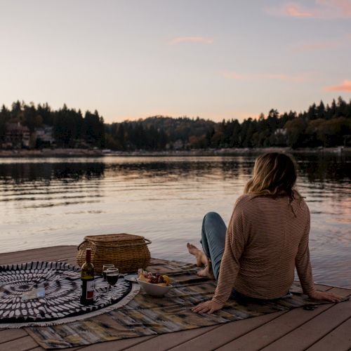 A woman relaxes on a lakeside deck with a picnic at sunset, watching calm water and trees turn golden.