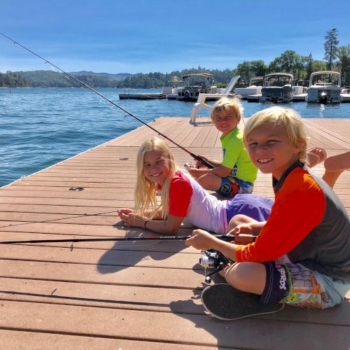 Three kids smiling on a dock by a lake, holding fishing rods with boats moored in the background.