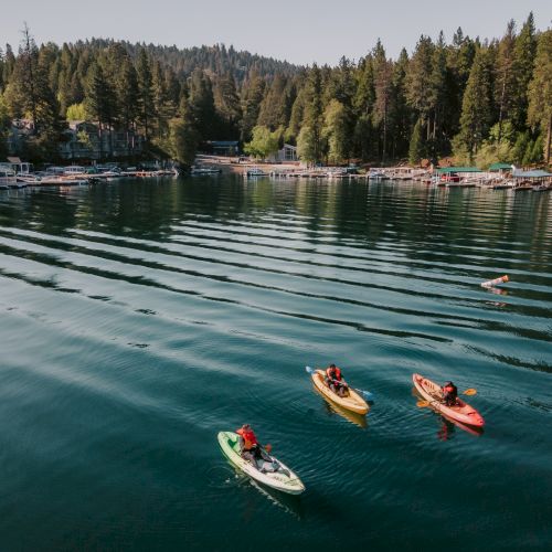 Kayakers paddle on a calm lake near a marina, with docks and boats beside a forested hillside of cabins and trees.