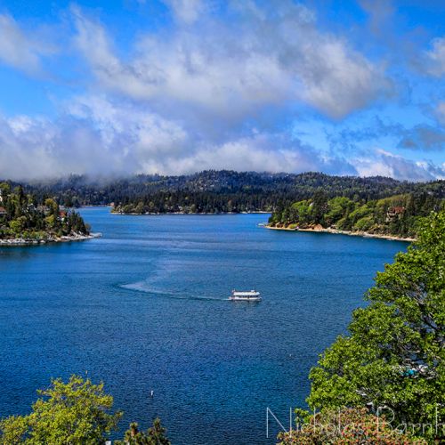 A scenic forested lake under dramatic blue clouds, with small islands and a distant boat drifting across the water.