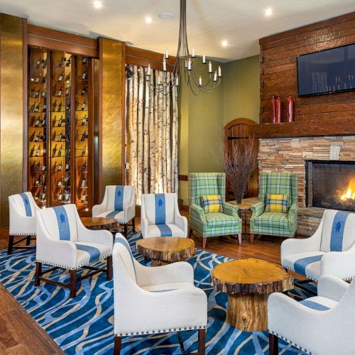 A cozy lounge with cushioned chairs, a stone fireplace, chandelier, wine bottle display wall, plaid armchairs, and patterned rug.