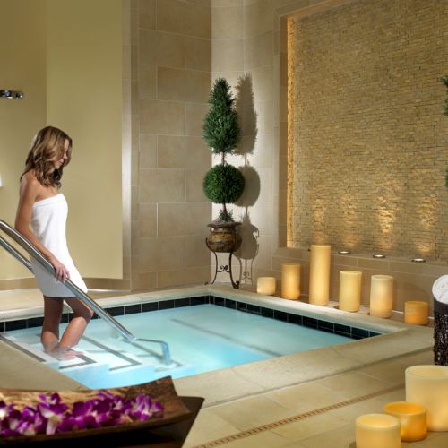 A woman steps into a tiled spa hot tub beside candles and potted evergreens, creating a relaxing wellness scene.