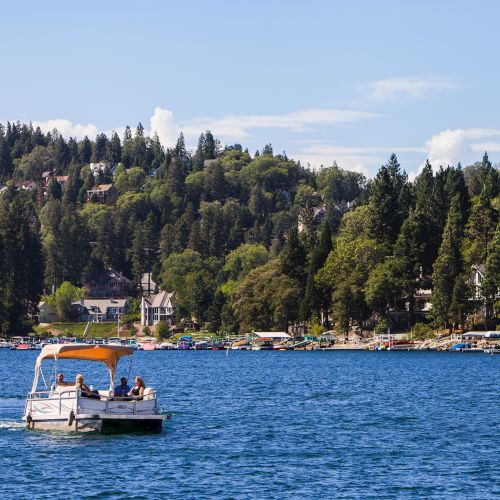 A sunny lakeside scene with a small boat cruising past a dock, surrounded by forested hills and hillside houses.