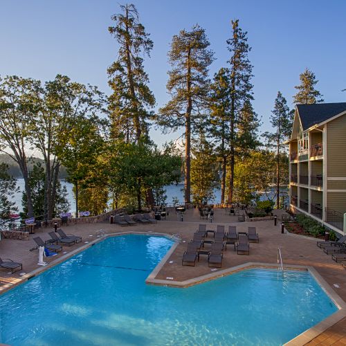 An outdoor hotel pool with lounge chairs by the water, surrounded by tall trees and a lakeside building with balconies.