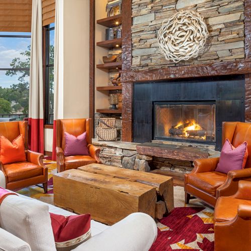 A cozy living room with a stone fireplace, leather chairs and colorful pillows, wooden shelves, and large windows by a lake view.