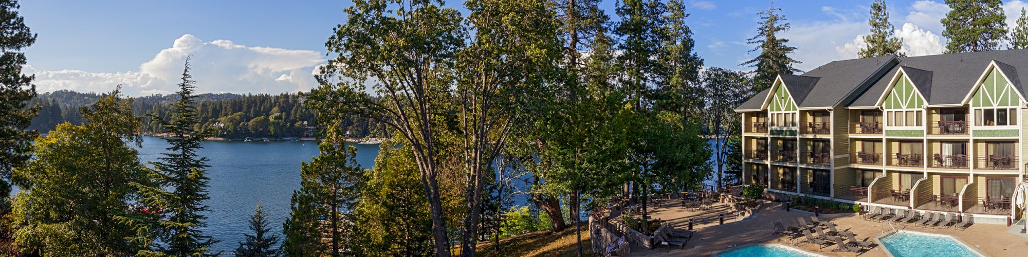Lakeside resort with a bright blue swimming pool, sun loungers, and multi-story cabins beside a calm lake and trees.