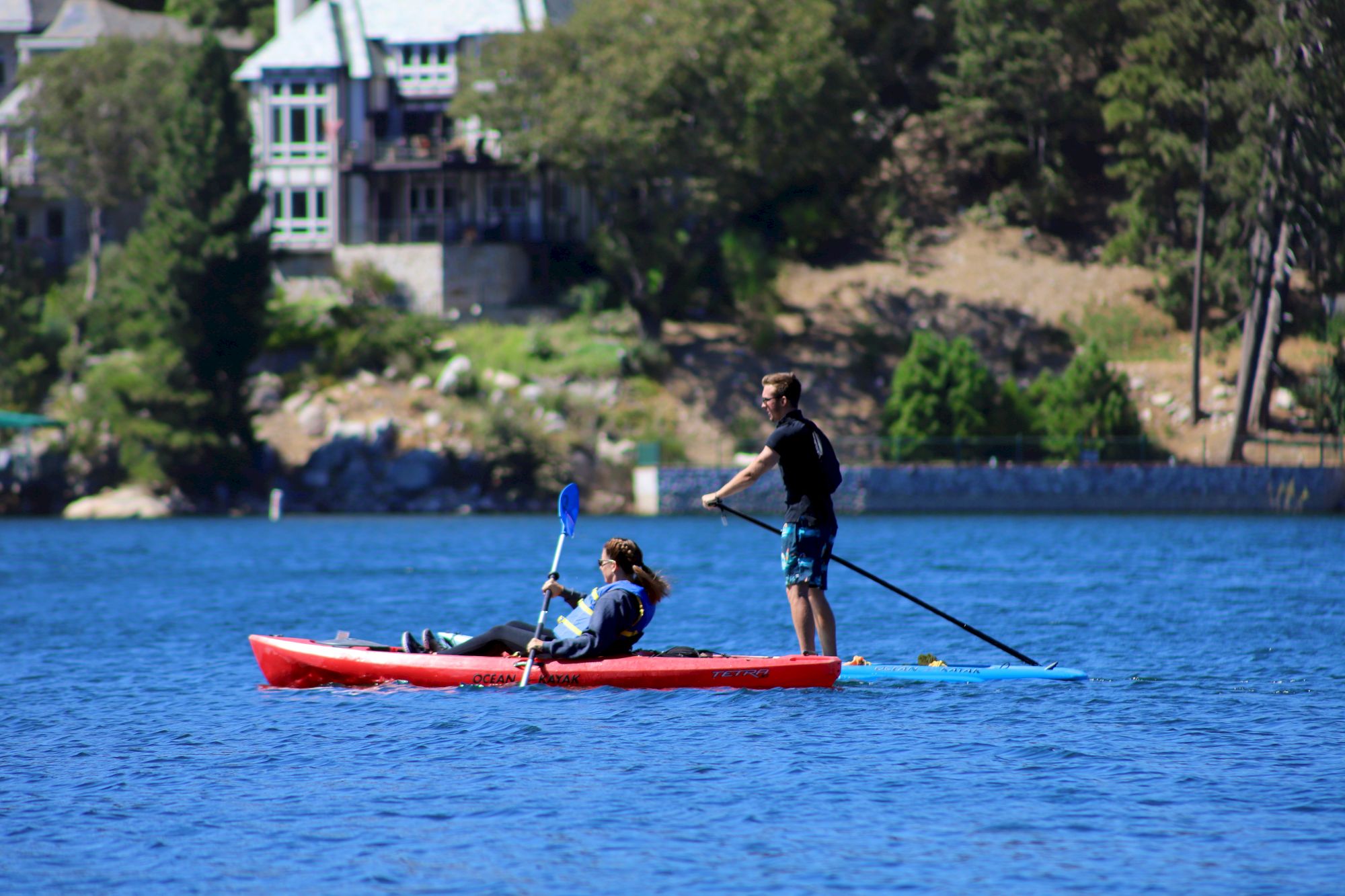 Two paddlers glide on a red kayak across a blue lake, with a forested shoreline and buildings in the background.