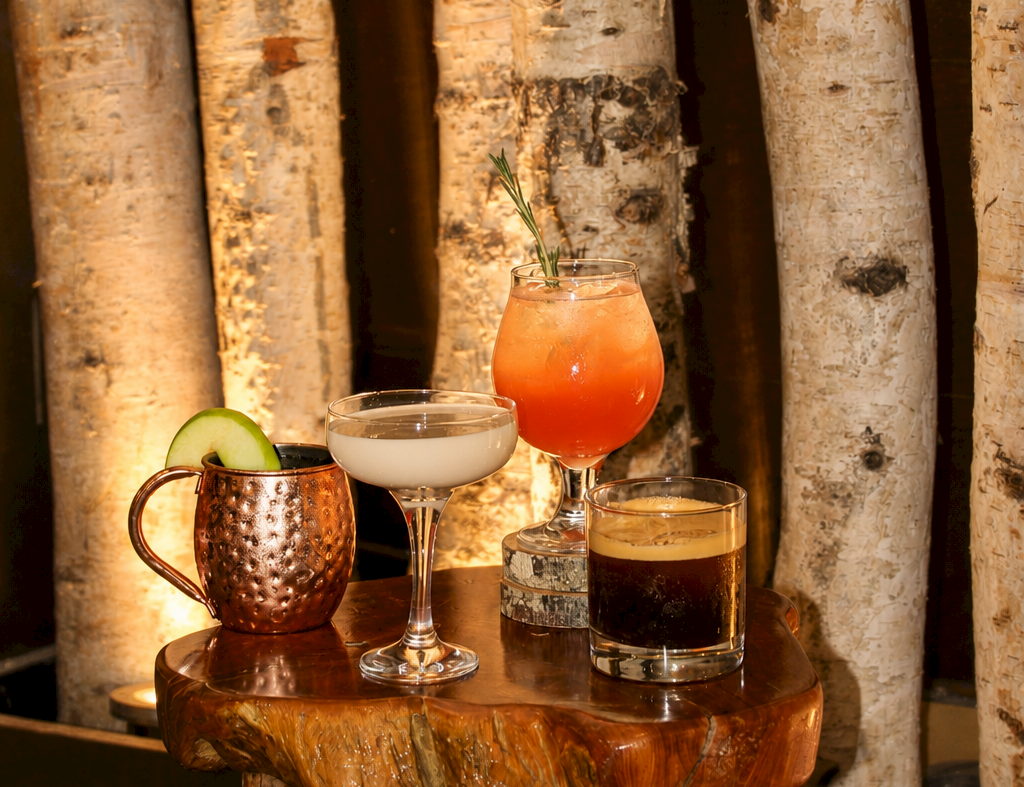 Four cocktails on a rustic wooden tray&mdash;orange, white, dark, and coffee&mdash;garnished with cucumber beside birch logs.