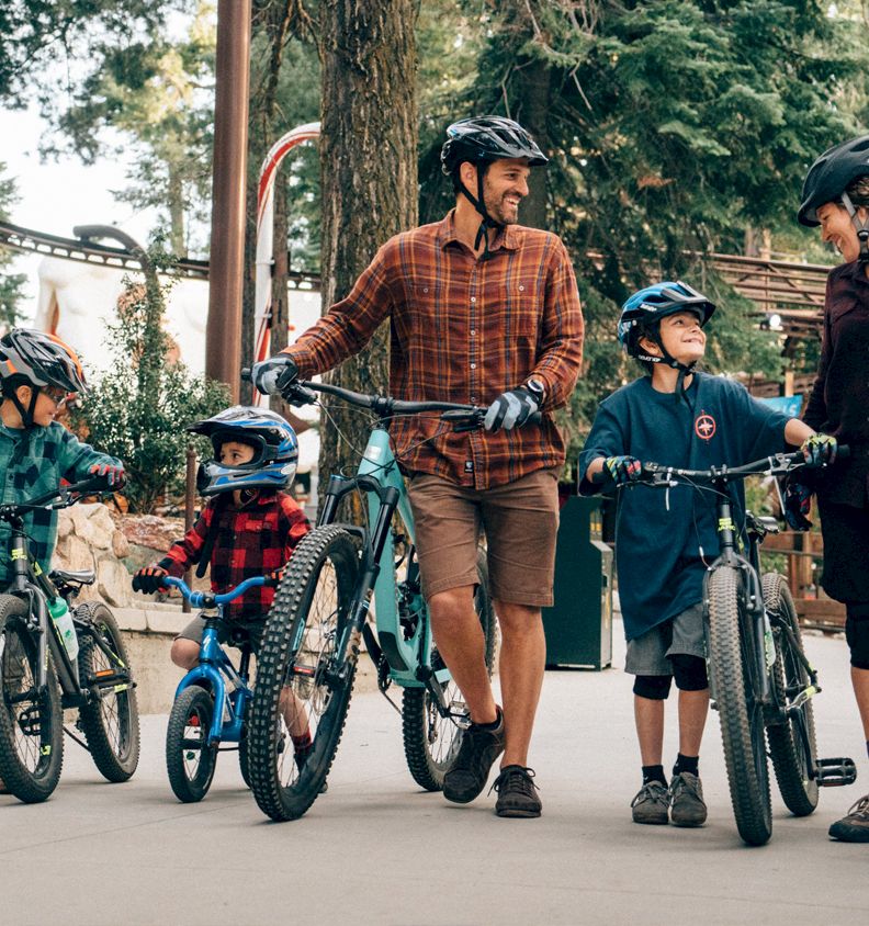 A family of cyclists in helmets rides and strolls together outdoors by the bikes in a forest park.