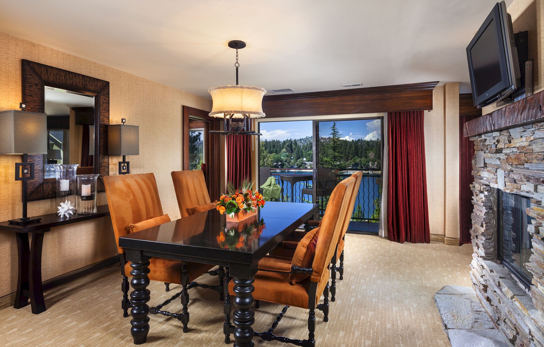 A furnished dining room with a chandelier, dark wood table and chairs, a TV, a stone fireplace wall, and a window view outside.