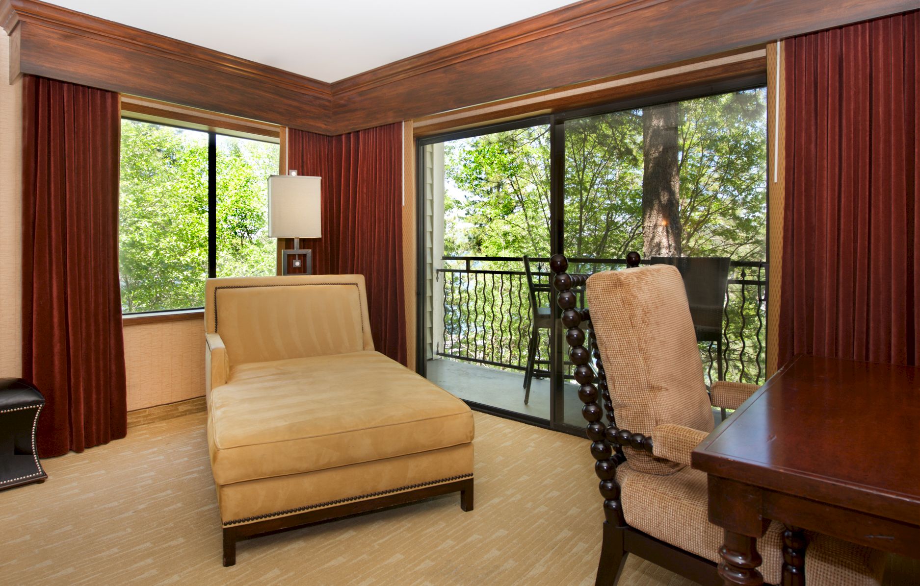 A cozy bedroom with a bed, red curtains, a lamp, and patio seating by large sliding doors overlooking green trees.