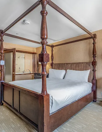 A spacious hotel bedroom with a dark four-poster bed, white bedding, nightstands, and warm wall lighting.