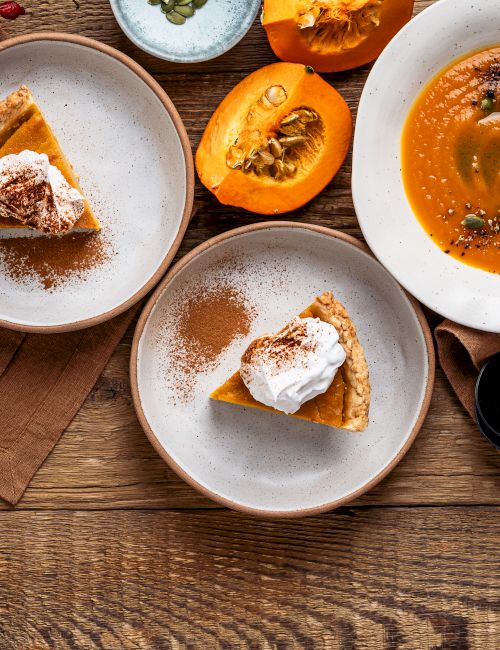 A rustic spread of pumpkin soup and pumpkin pie slices, topped with cream, seeds, and herbs on wooden boards.