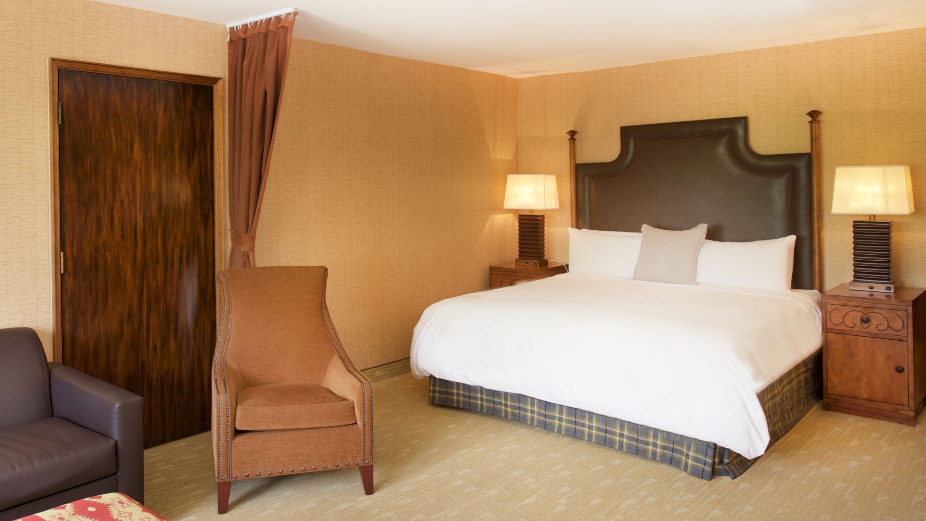 A hotel-style bedroom with a large bed, nightstands and lamps, a brown chair, and a wooden door with curtains.