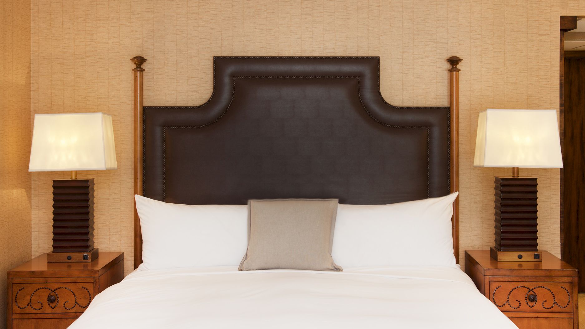 A styled bedroom with a large bed, dark tufted headboard, two table lamps, and wooden nightstands against a beige wall.