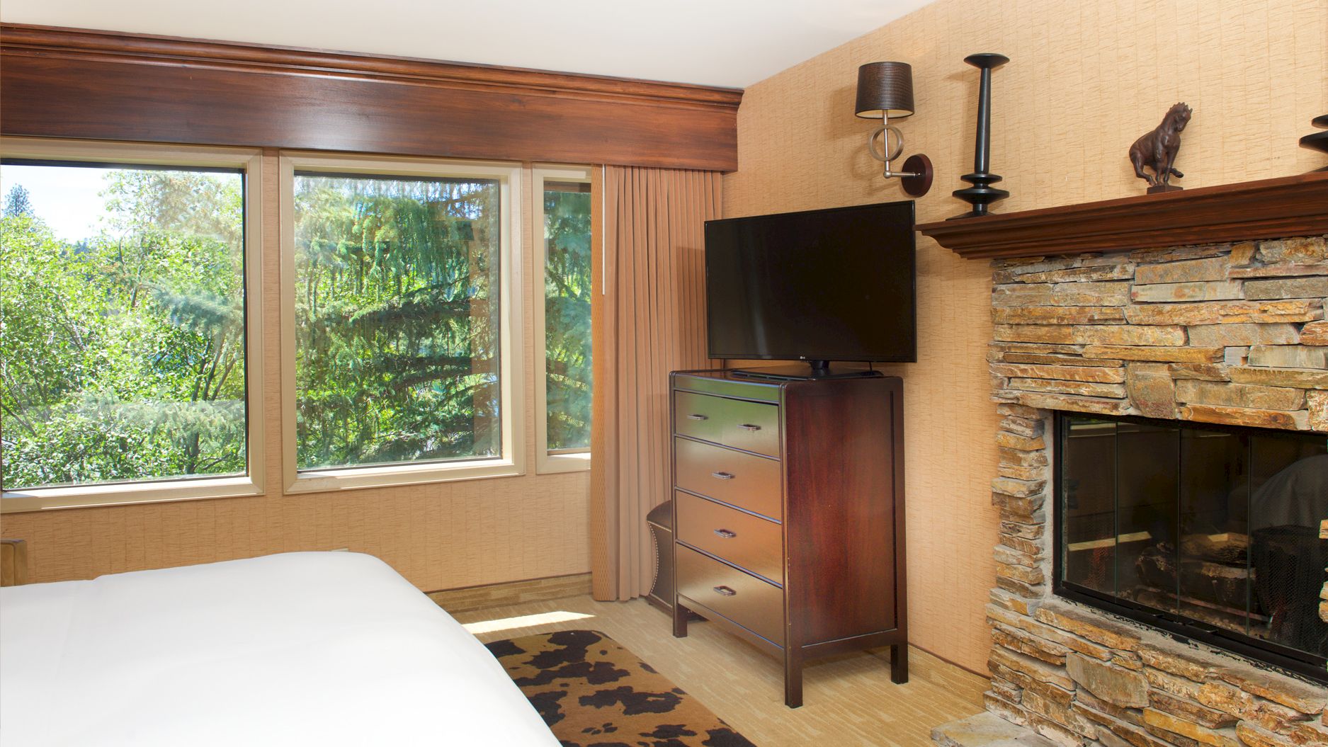 A modern bedroom with large windows, a TV on a dresser, and a stone fireplace with wall sconces.