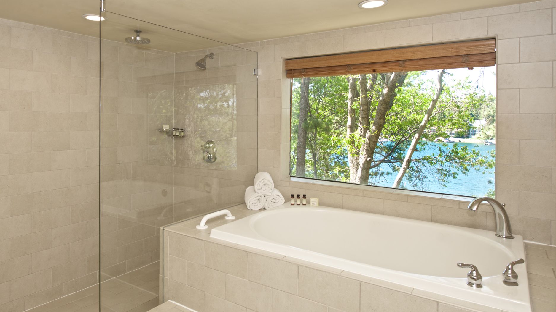 A bright bathroom with a bathtub, glass shower, tiled walls, and a window view of trees and water.