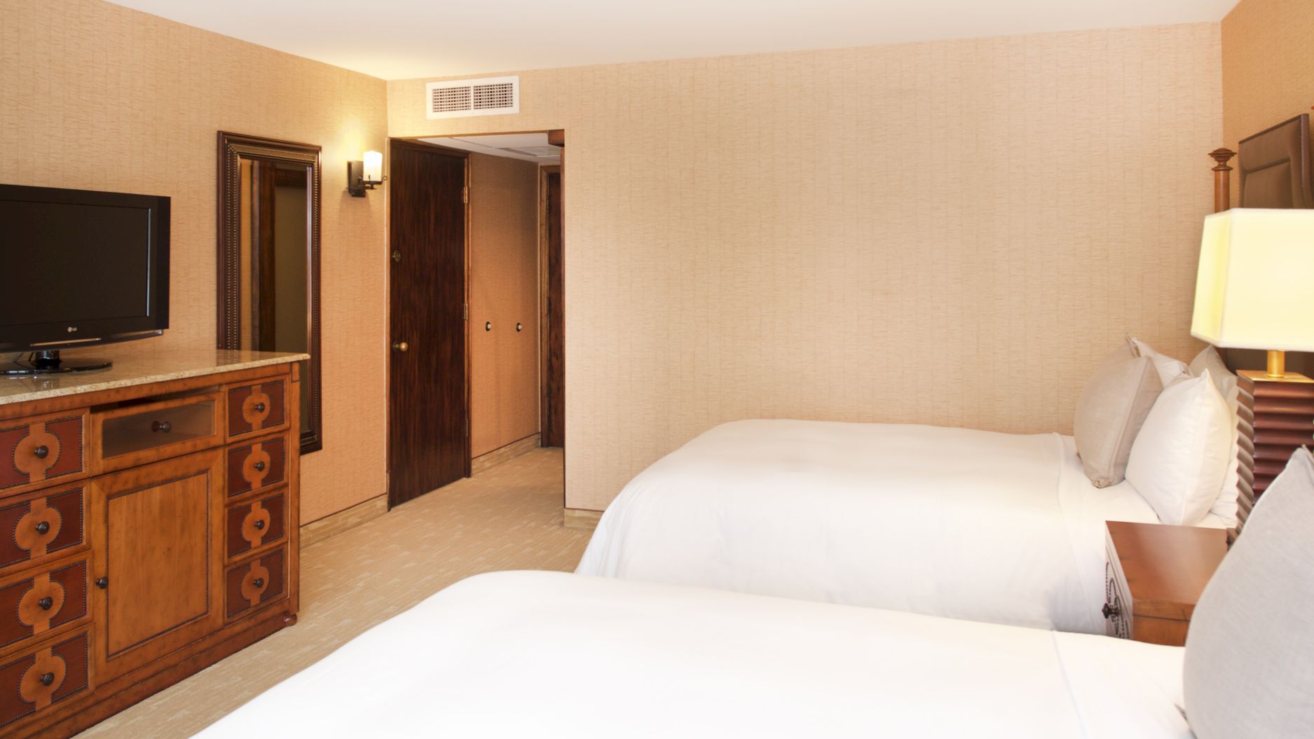 A tidy hotel room with two beds, a dresser with TV, a lamp, and doors in a beige, textured wall.