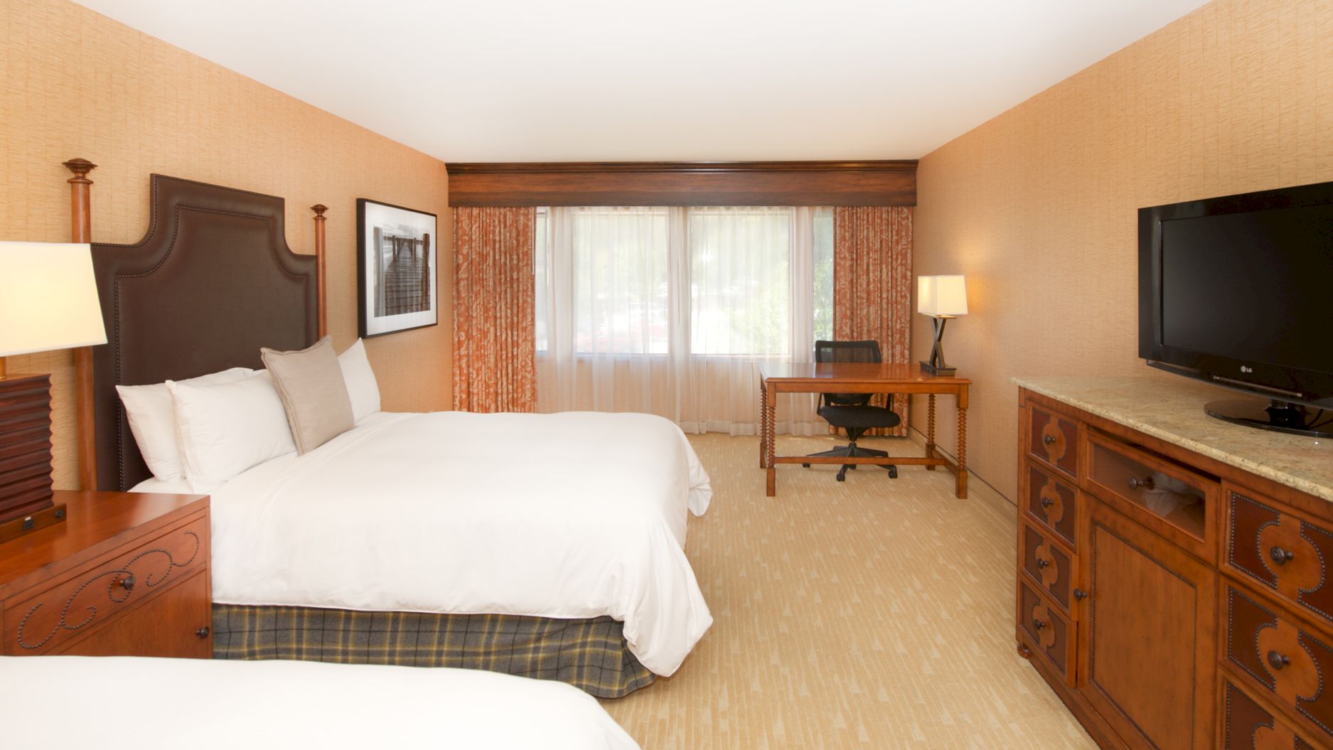 A hotel room with two beds, a desk and lamp, and a flat-screen TV by the window with curtains.