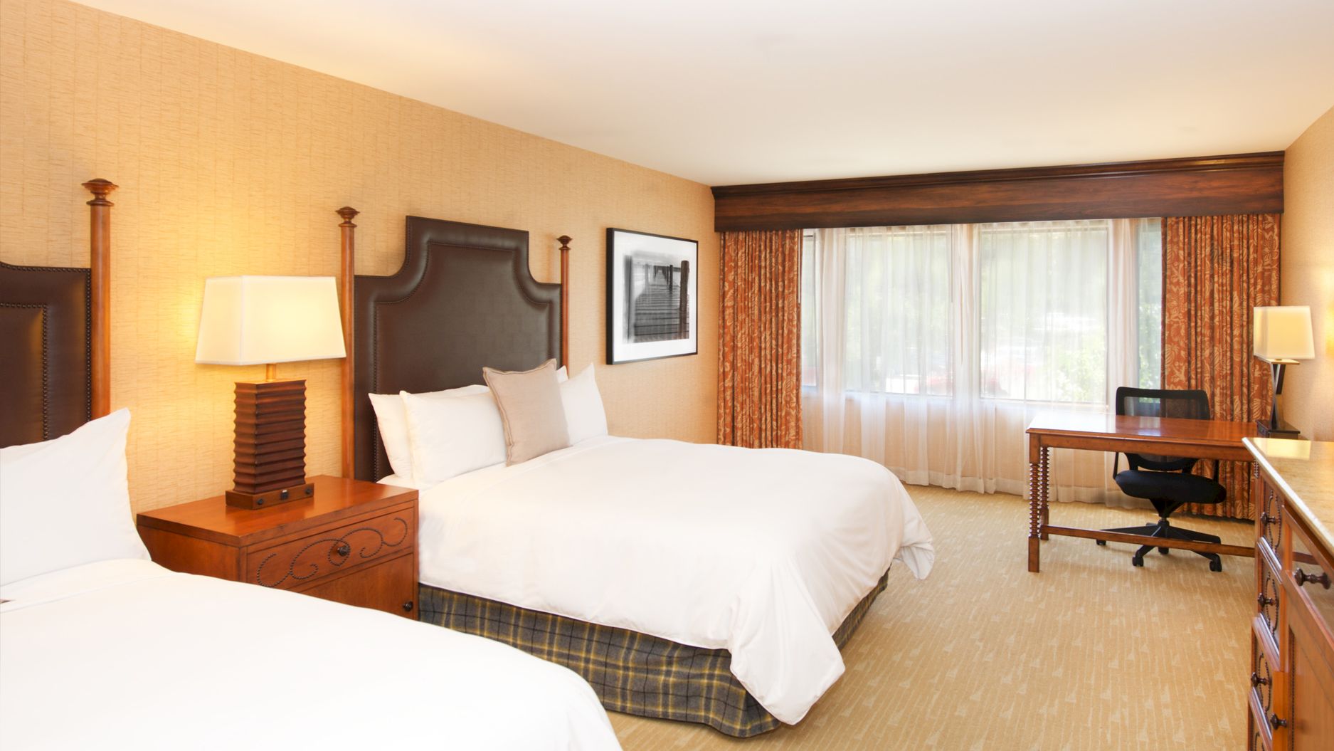 A hotel-style bedroom with two beds, bedside lamps, a desk and chair, and orange curtains by a window.