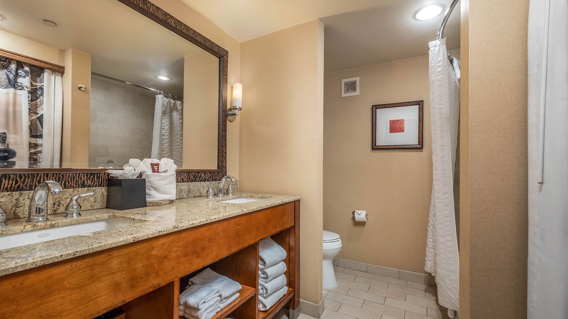 A hotel bathroom with granite vanity, mirror, towels, toilet, and a glass-enclosed shower with a showerhead.