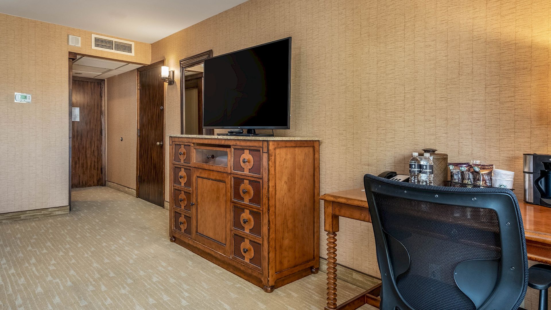 Hotel room corner with a wall-mounted TV, wooden cabinet, desk setup, bottled drinks, and a blue office chair by the wall.