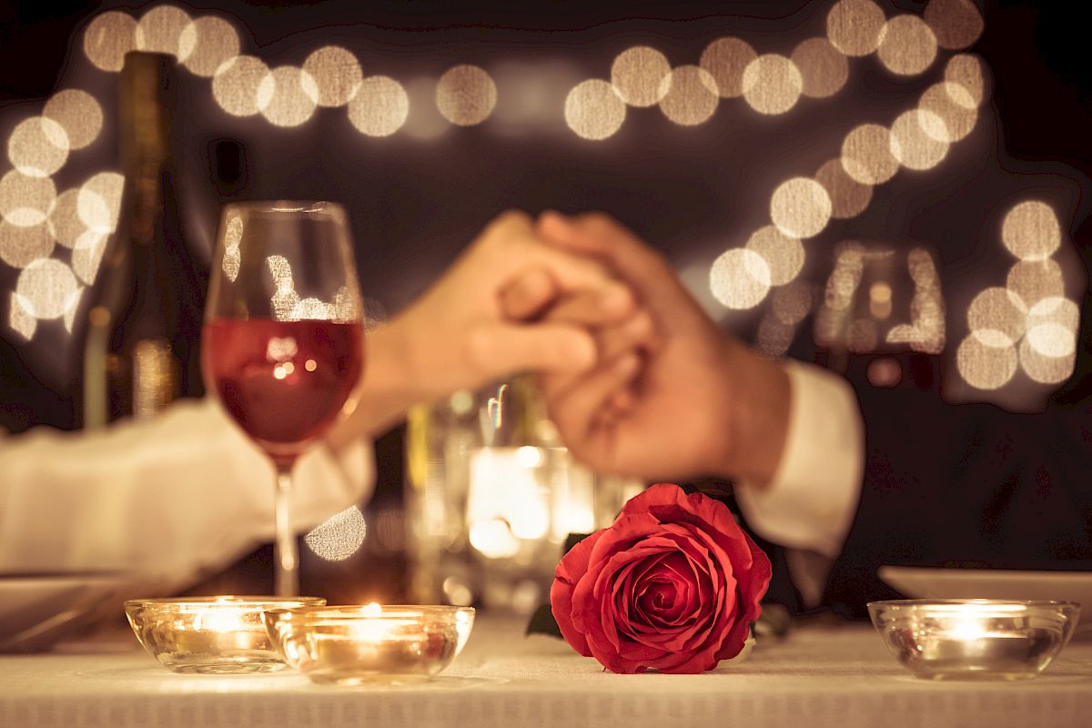 A romantic dinner setup with a couple holding hands, wine glasses, candles, a red rose, and blurred fairy lights in the background.