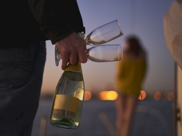 Two people walking toward a beach at nighttime, one of which is holding a bottle of sparkling wine and two champagne flutes.