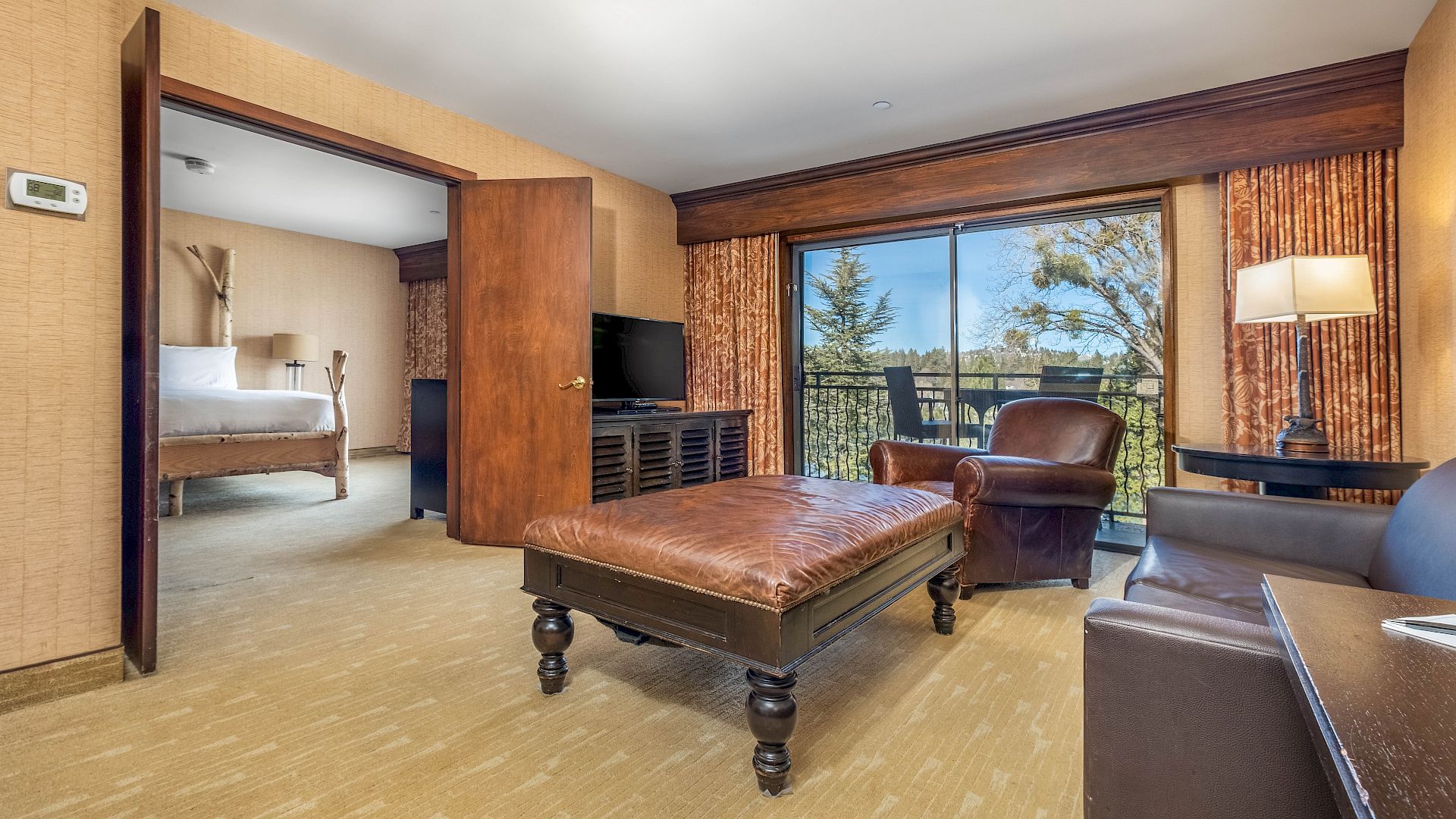 A cozy hotel room/suite with a leather bed, TV, armchairs, and large windows overlooking a garden.