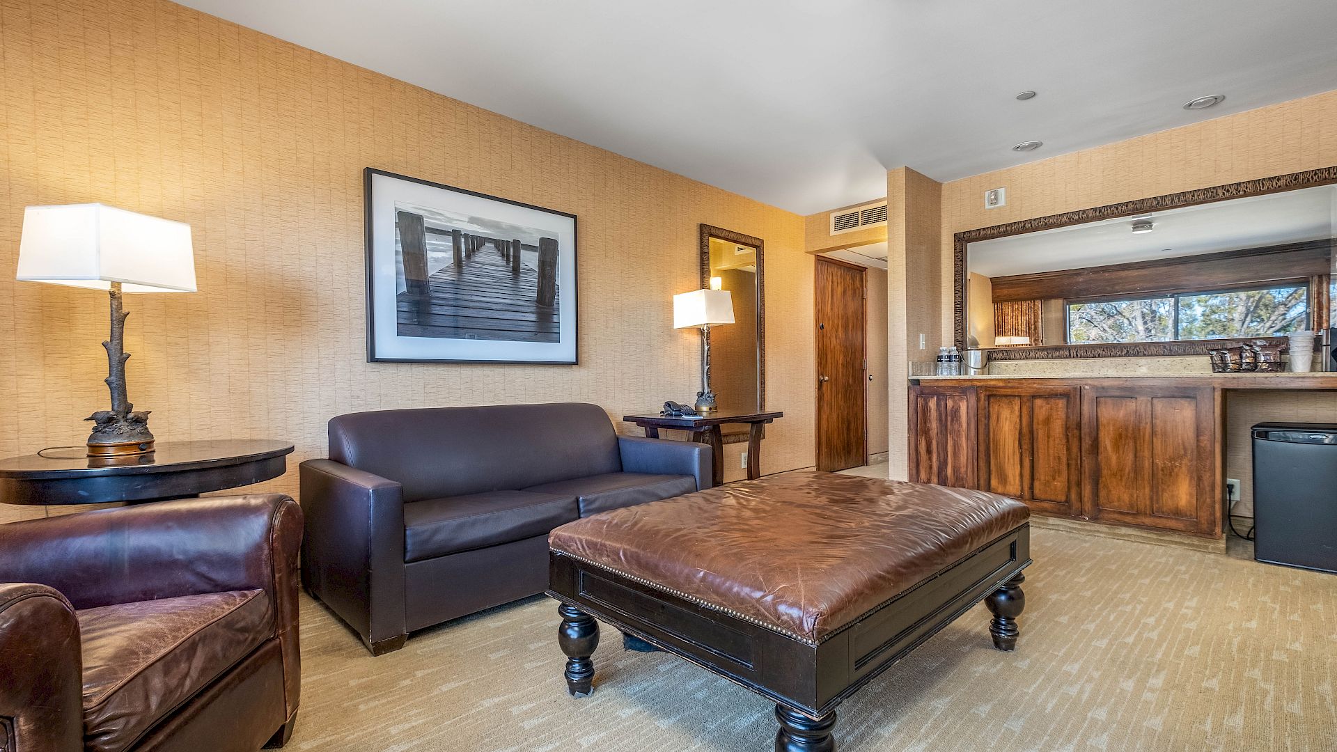 A cozy hotel-style room with brown sofas, a leather ottoman, lamps, framed wall art, and a wooden bar counter.