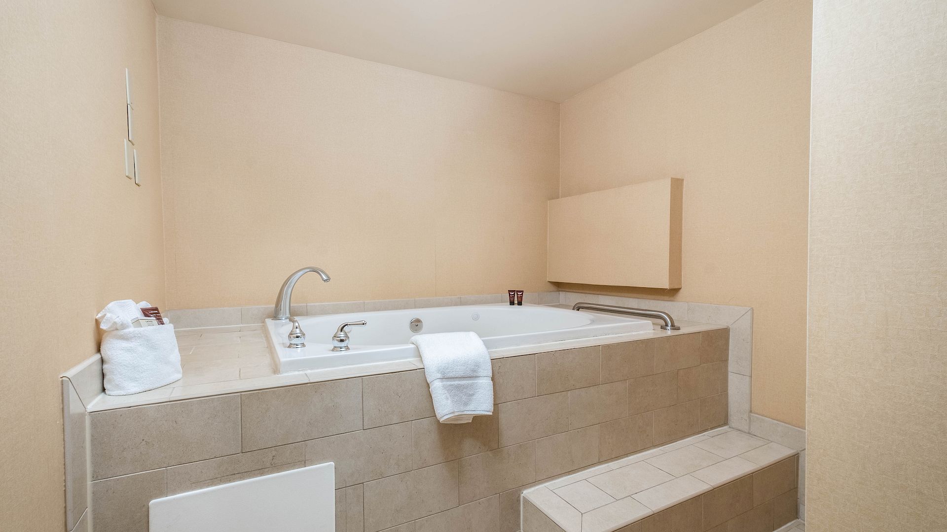 A hotel bathroom with a long white bathtub, towels, and faucets set in beige tile walls.