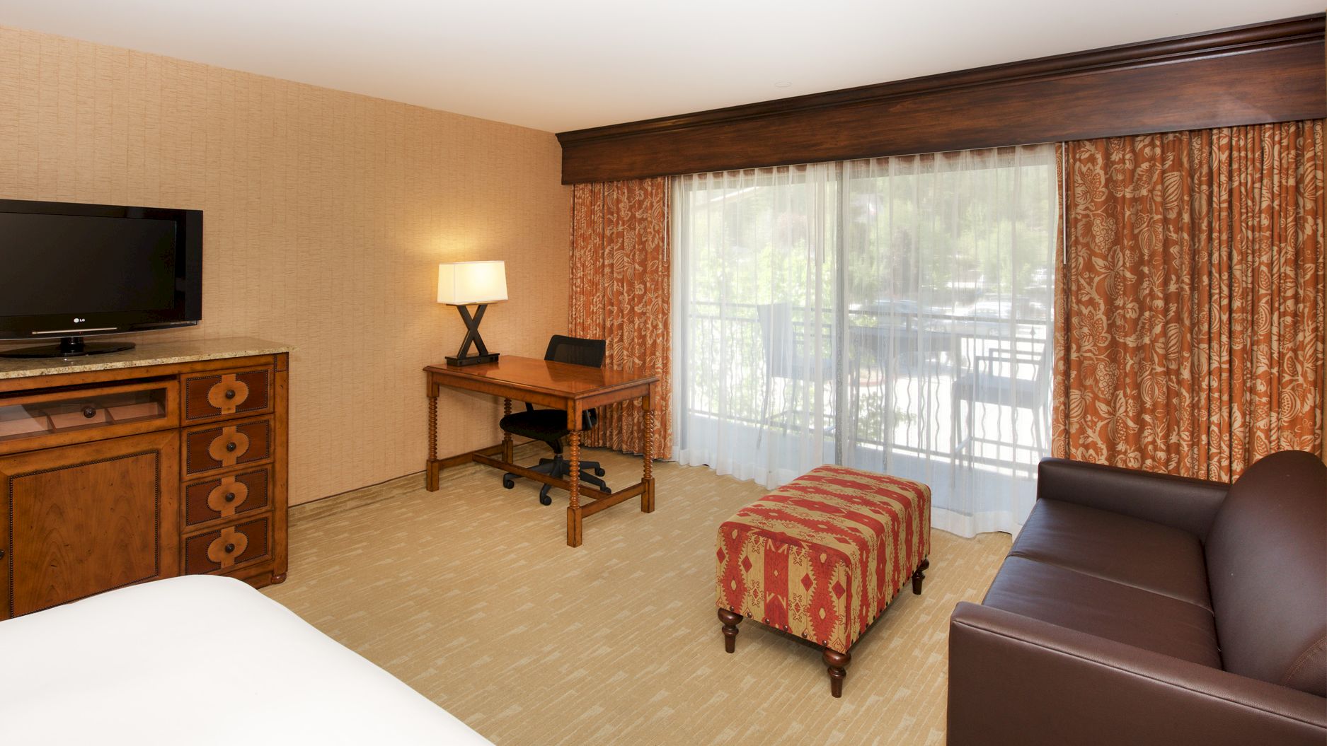 A cozy bedroom-office with a desk, lamp, TV on a dresser, bed with patterned blanket, and large curtained window.