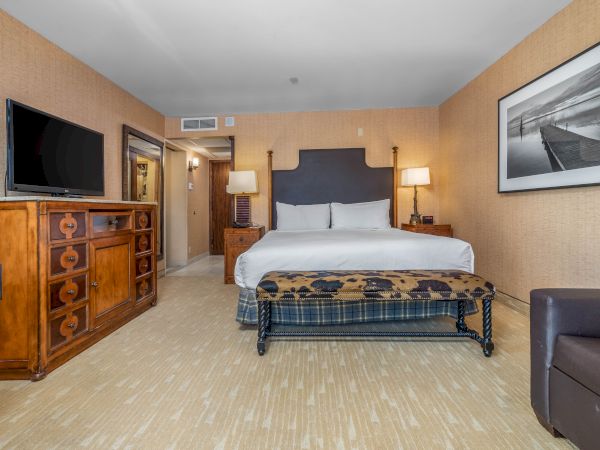 A hotel room with a king-size bed, TV on a dresser, lamps, framed wall art, and beige walls.