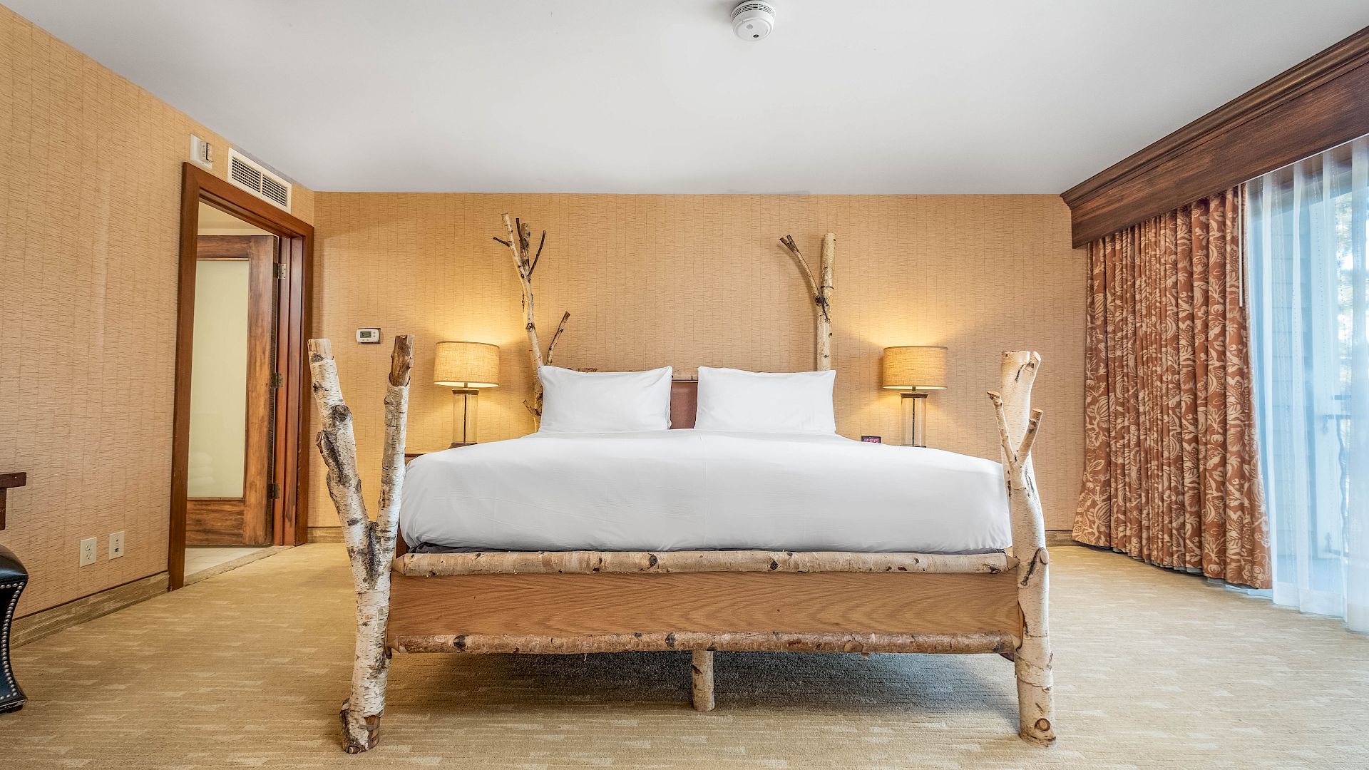 A cozy hotel bedroom with a log-framed bed, two white pillows, bedside lamps, birch decor, and curtained window.