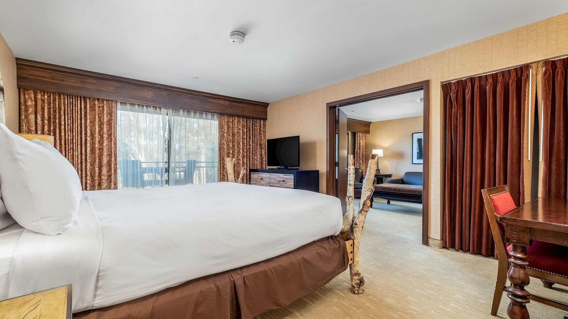 A hotel room with a large bed, patterned curtains, a TV on a stand, and an adjoining seating area.