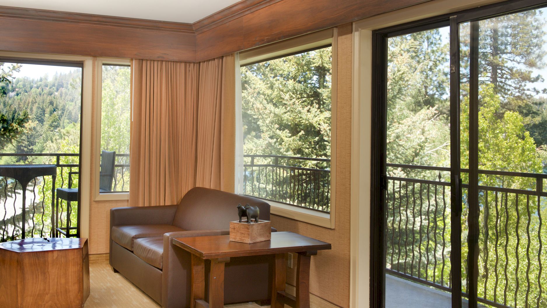 Bright lounge nook with a sofa, wooden table with decorative animal figures, and large windows overlooking a lush green forest.